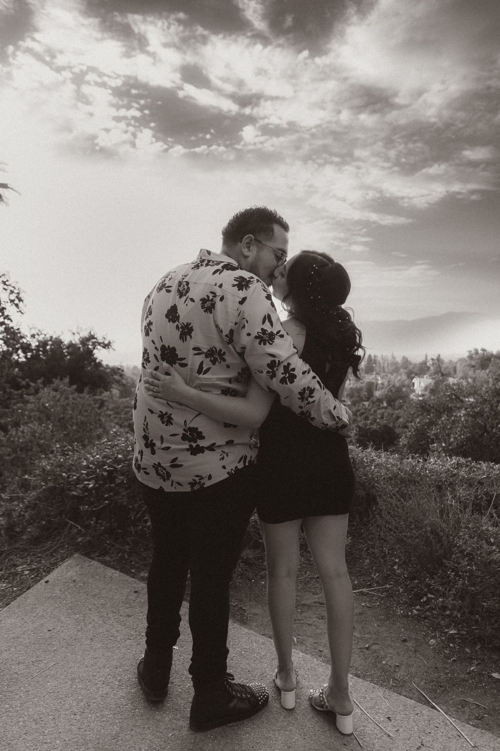 A romantic couple shares an intimate moment on a path with dramatic clouds in the background.