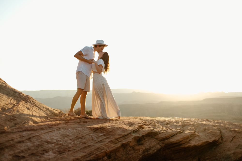 Romantic couple in white outfits embracing on desert canyon cliff edge during golden hour.