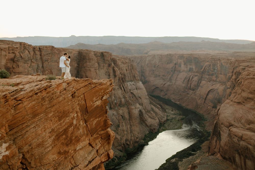 Two figures in white standing on dramatic cliff edge overlooking winding river canyon below.