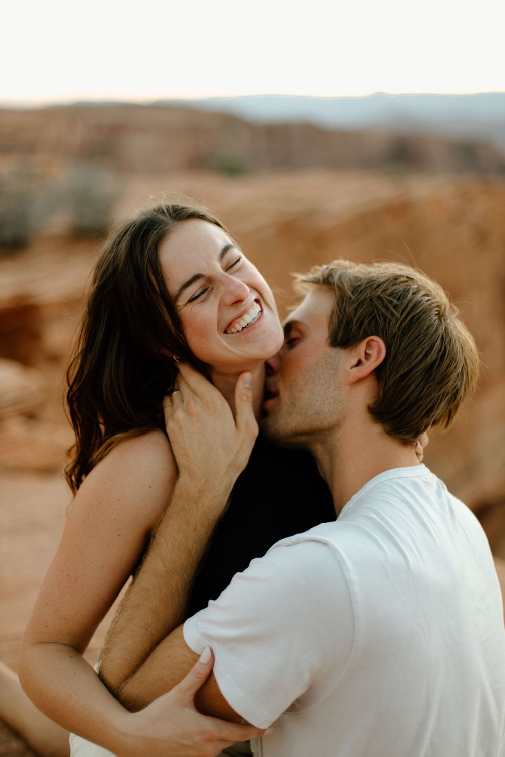 Joyful couple sharing intimate laughing moment against desert canyon backdrop at sunset.