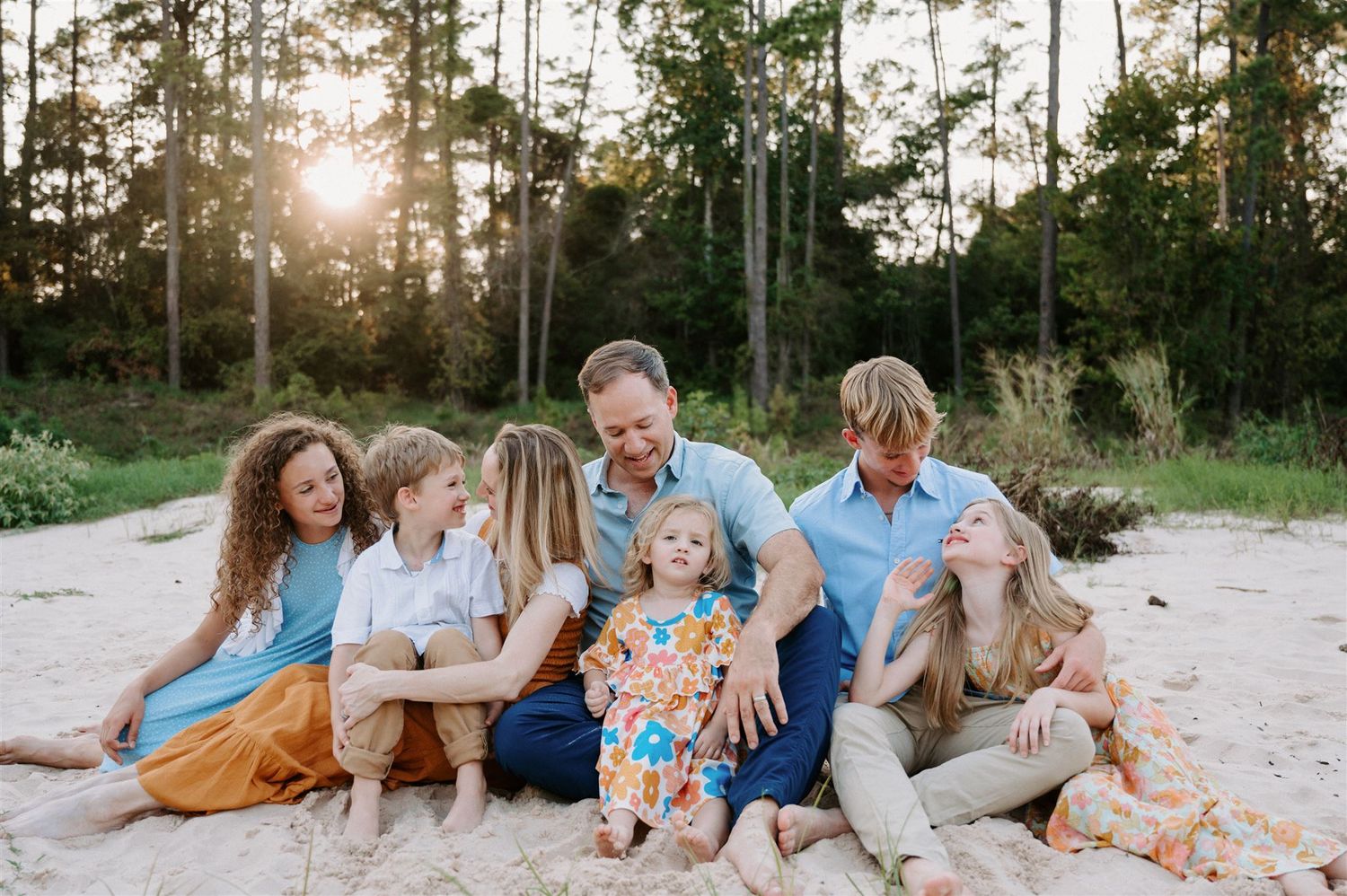 A cheerful family group shares laughter while sitting together on sandy ground at sunset.