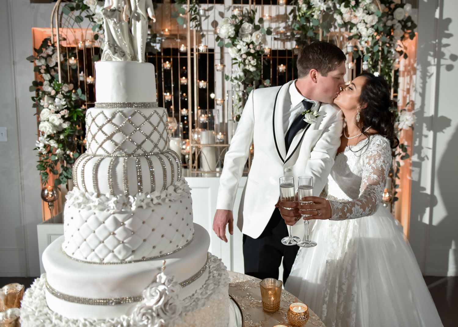 Elegant multi-tiered white wedding cake with geometric pattern sits beside couple sharing intimate moment.
