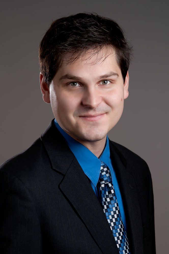 Young man with dark hair in black suit and blue checkered tie against gray background.