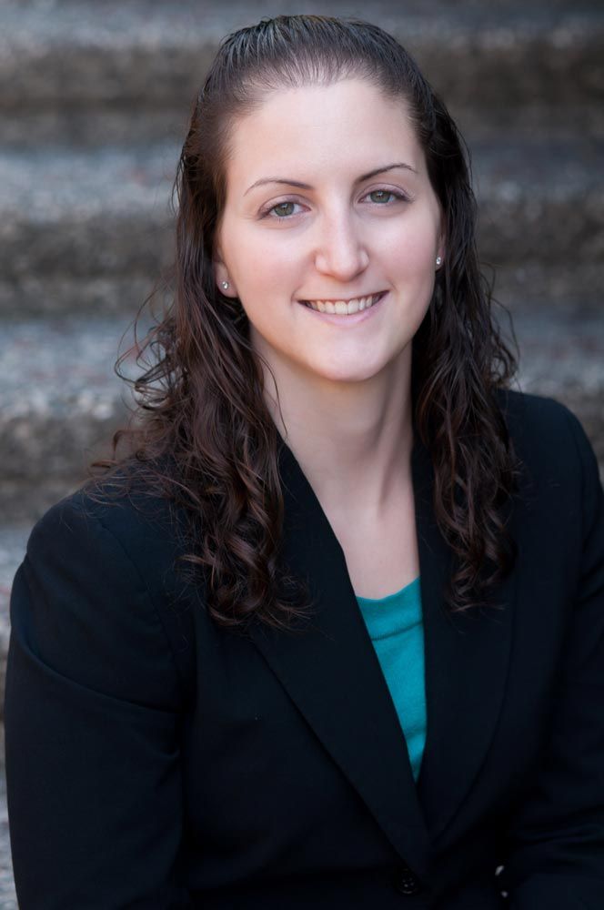 Young woman with curly dark hair in black blazer and teal top smiling on outdoor steps.