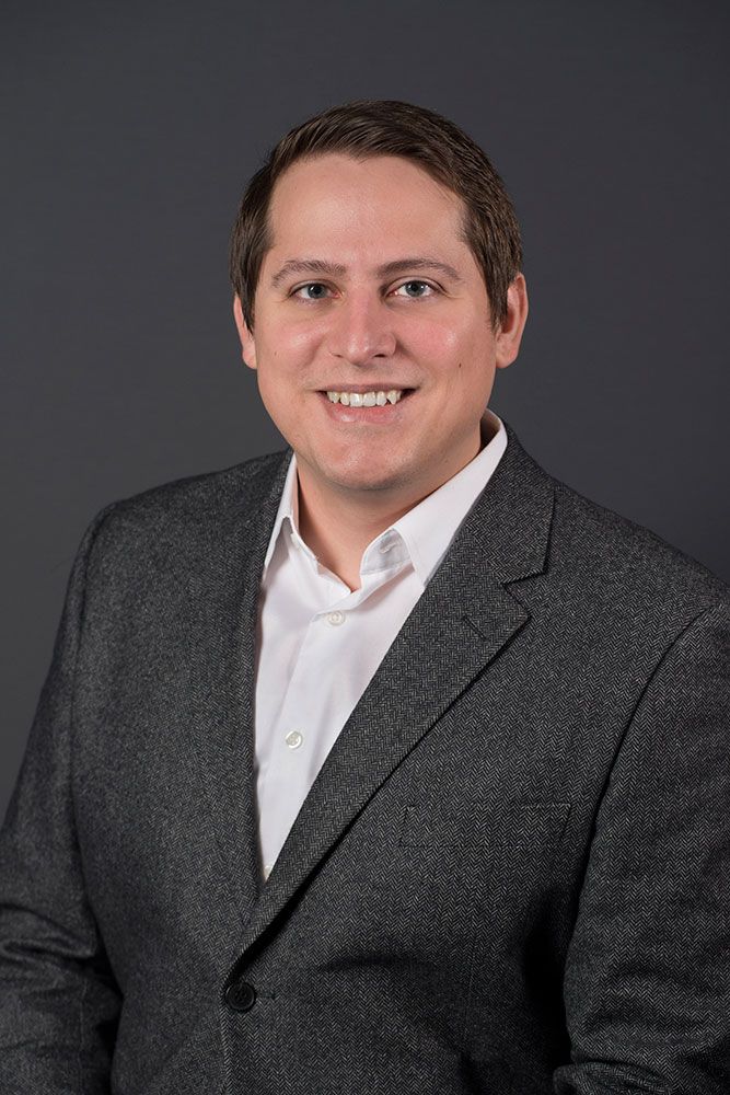 Smiling professional man with dark hair wearing a gray herringbone blazer and white shirt against a gray background.