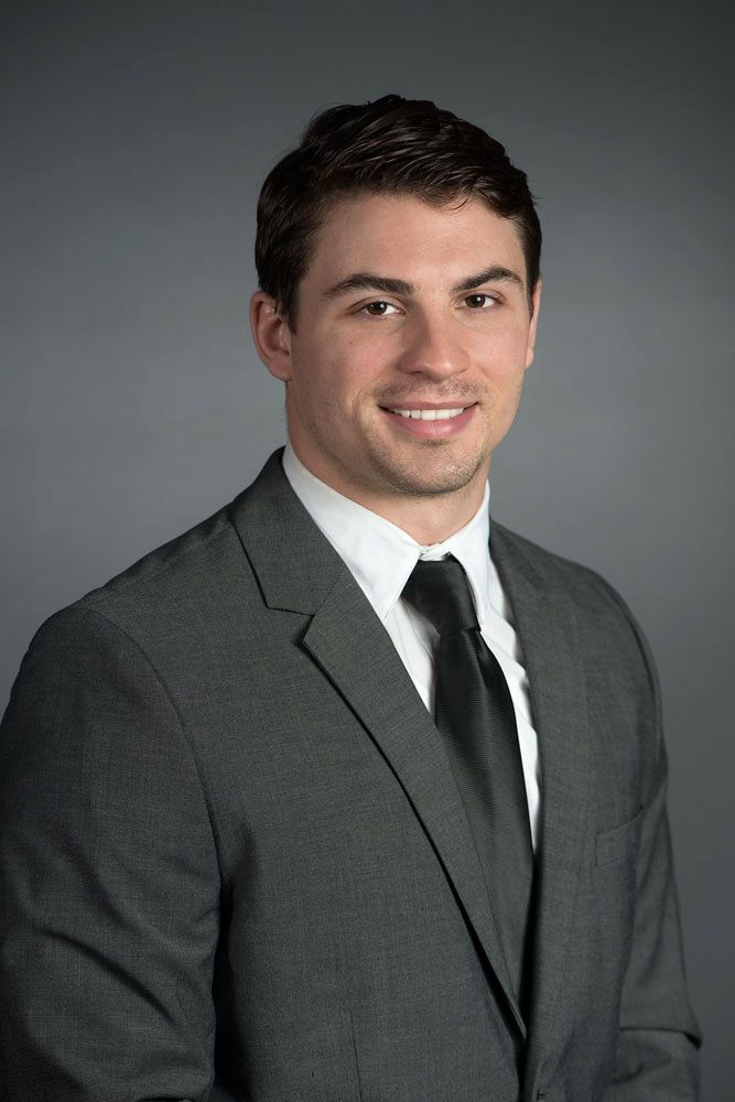 Smiling young professional man with dark hair wearing a gray suit and black tie against a gray background.