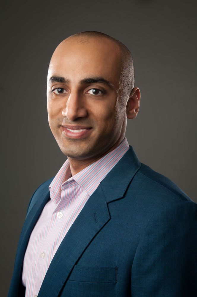 Bald man in teal blazer and pink striped shirt smiling professionally against gray background.