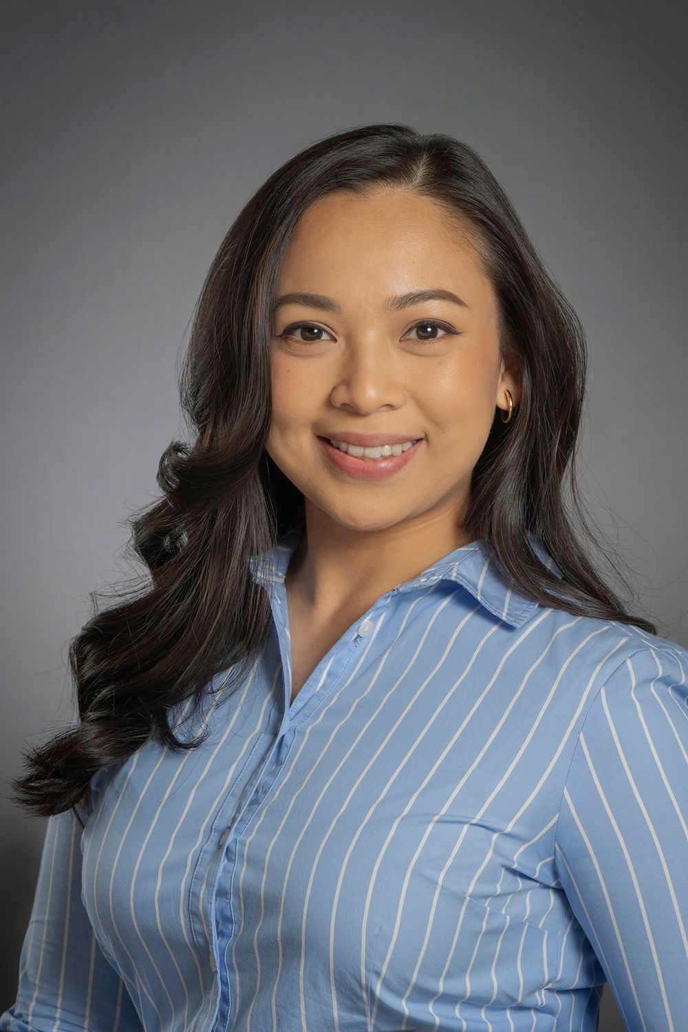 Smiling young woman with dark wavy hair in a blue striped shirt poses for a headshot.