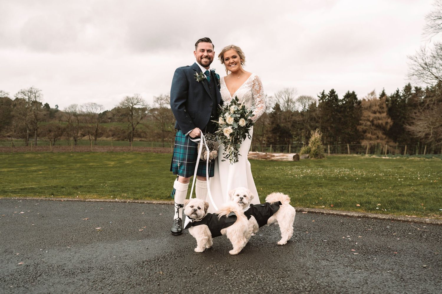 Couple in wedding attire with two white dogs posing outdoors in Scottish countryside.