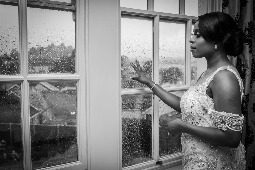 A woman in an elegant white dress gazes thoughtfully through a windowpane in black and white.