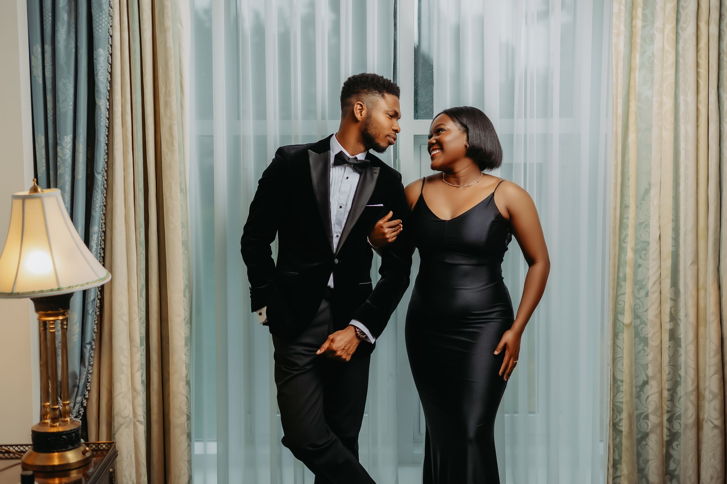 Elegant couple in formal evening wear pose together near window.