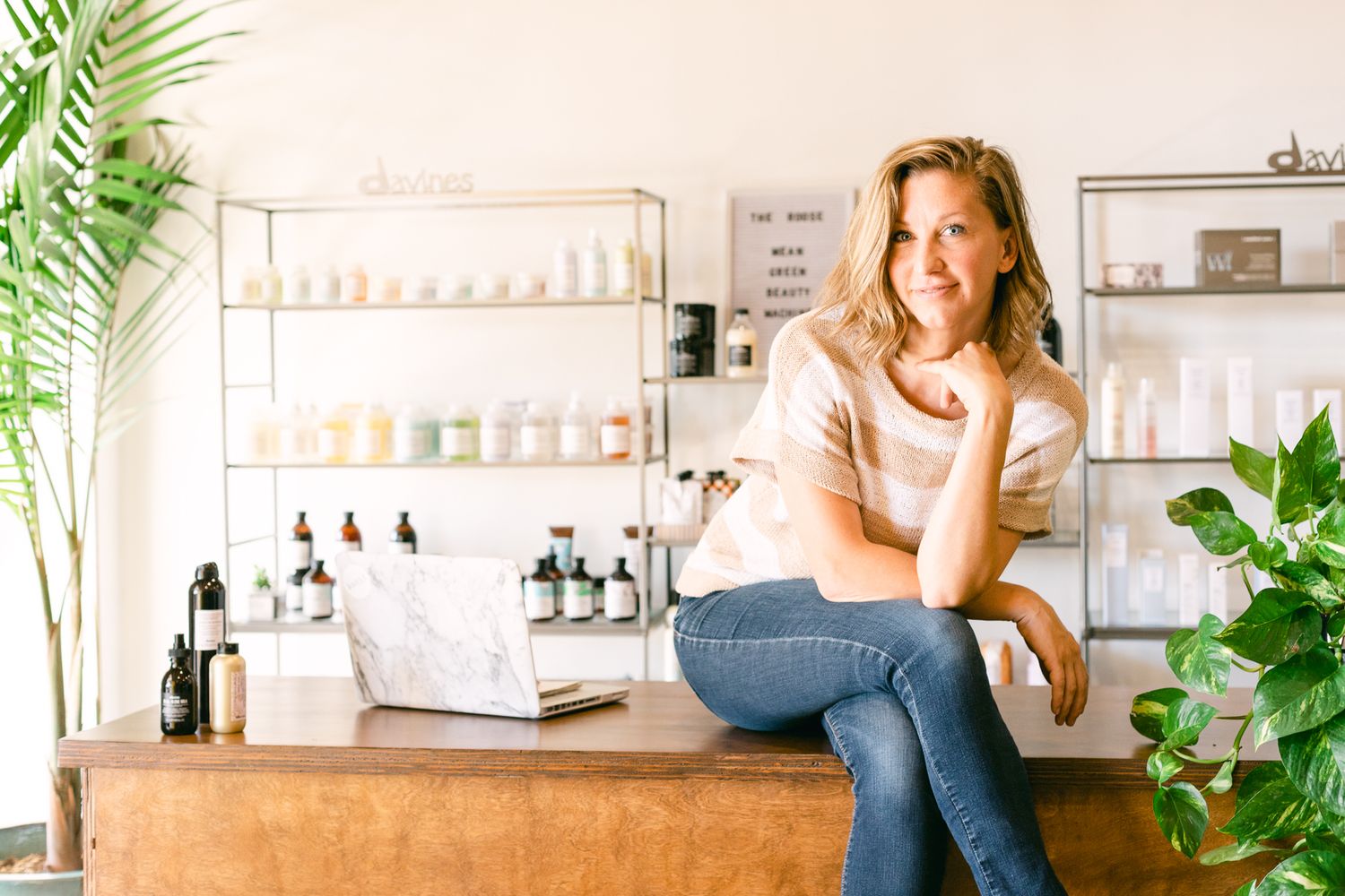 A blonde woman sits on a counter. She's in a striped shirt and there are hair products on shelves in the background.