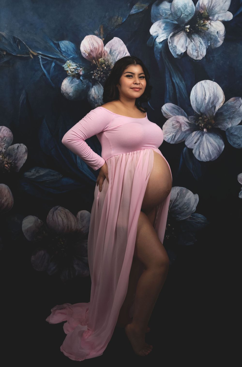 Maternity photo featuring a pink flowing dress against a dark backdrop with blue floral designs.