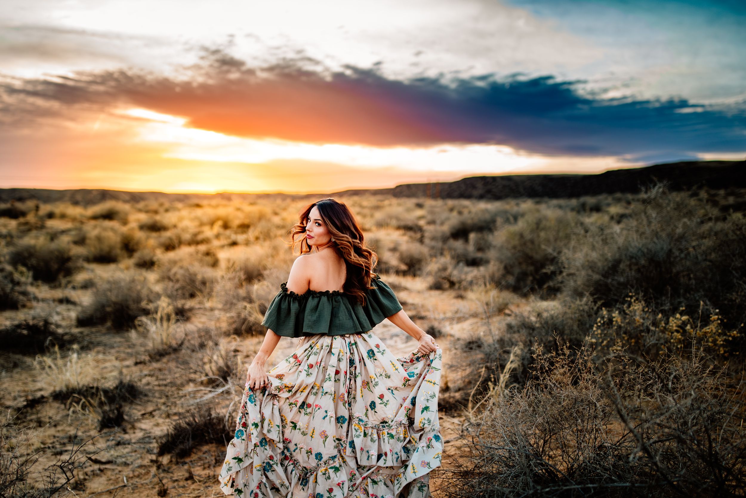 Flowing dress catches the wind against a desert sunset landscape with dramatic clouds.