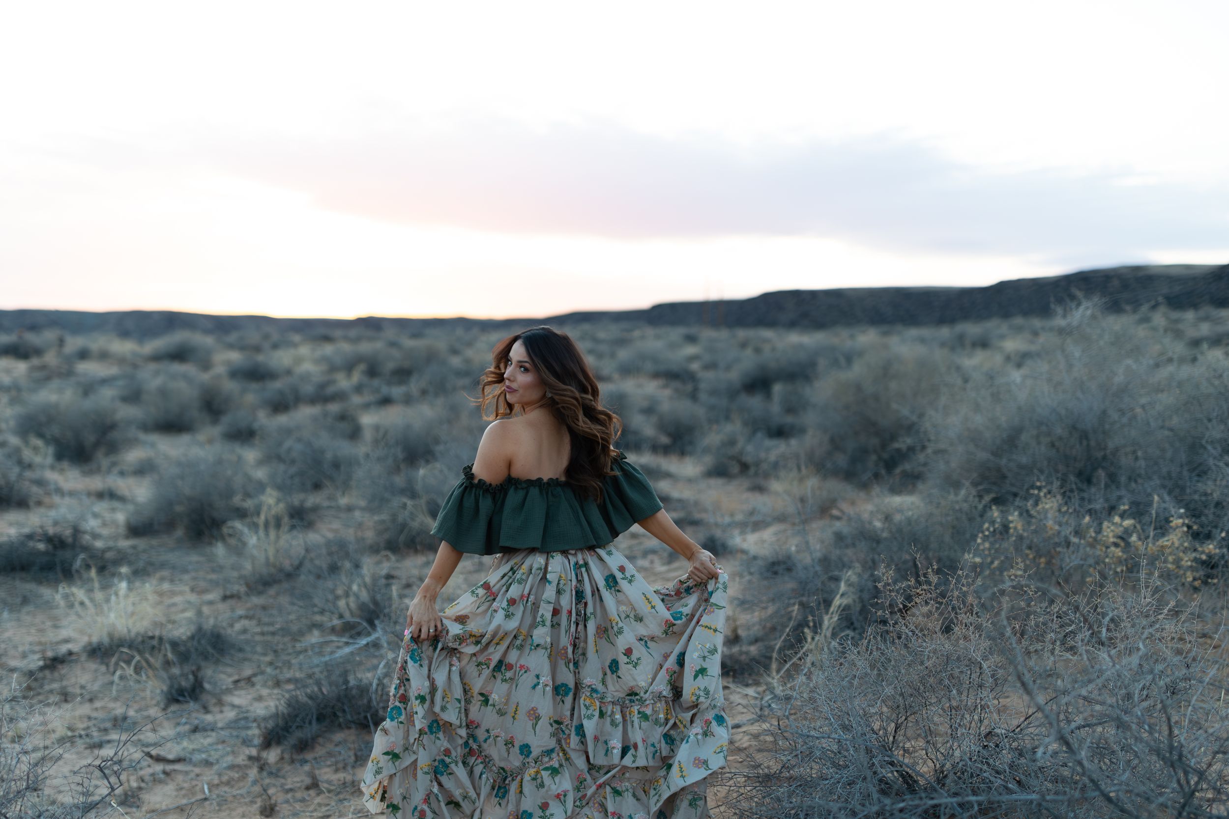 Desert landscape view with figure in flowing patterned dress against muted evening sky.