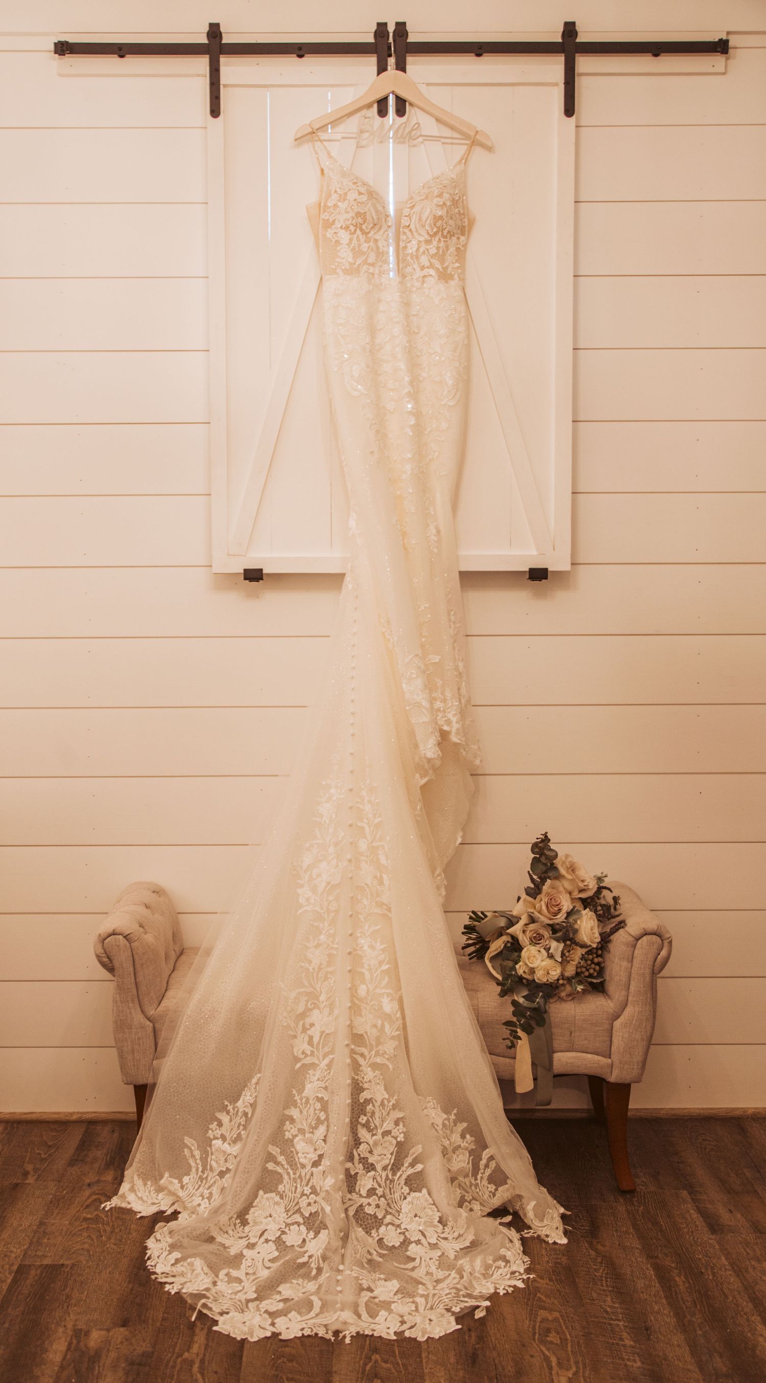 Elegant lace wedding dress hanging on barn door with floral bouquet below.