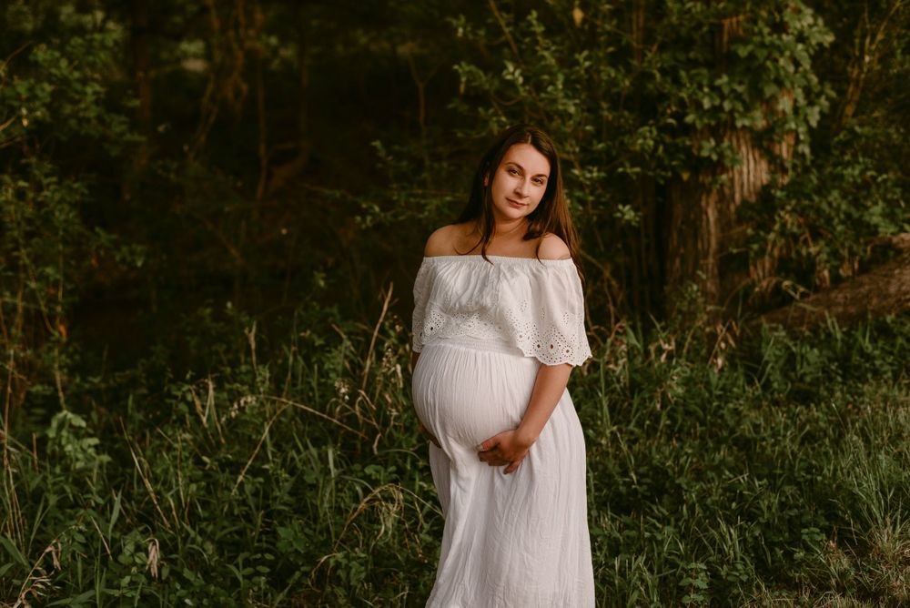Dreamy maternity portrait in white off-shoulder dress against lush forest backdrop.