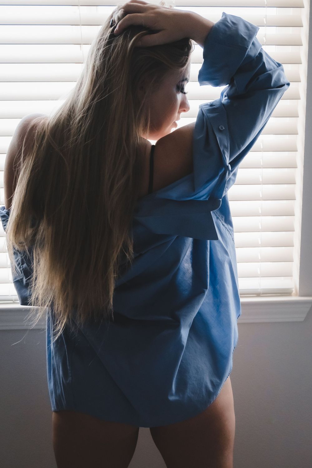 Young woman with long blonde hair posing in a blue oversized shirt near window blinds in soft natural light.