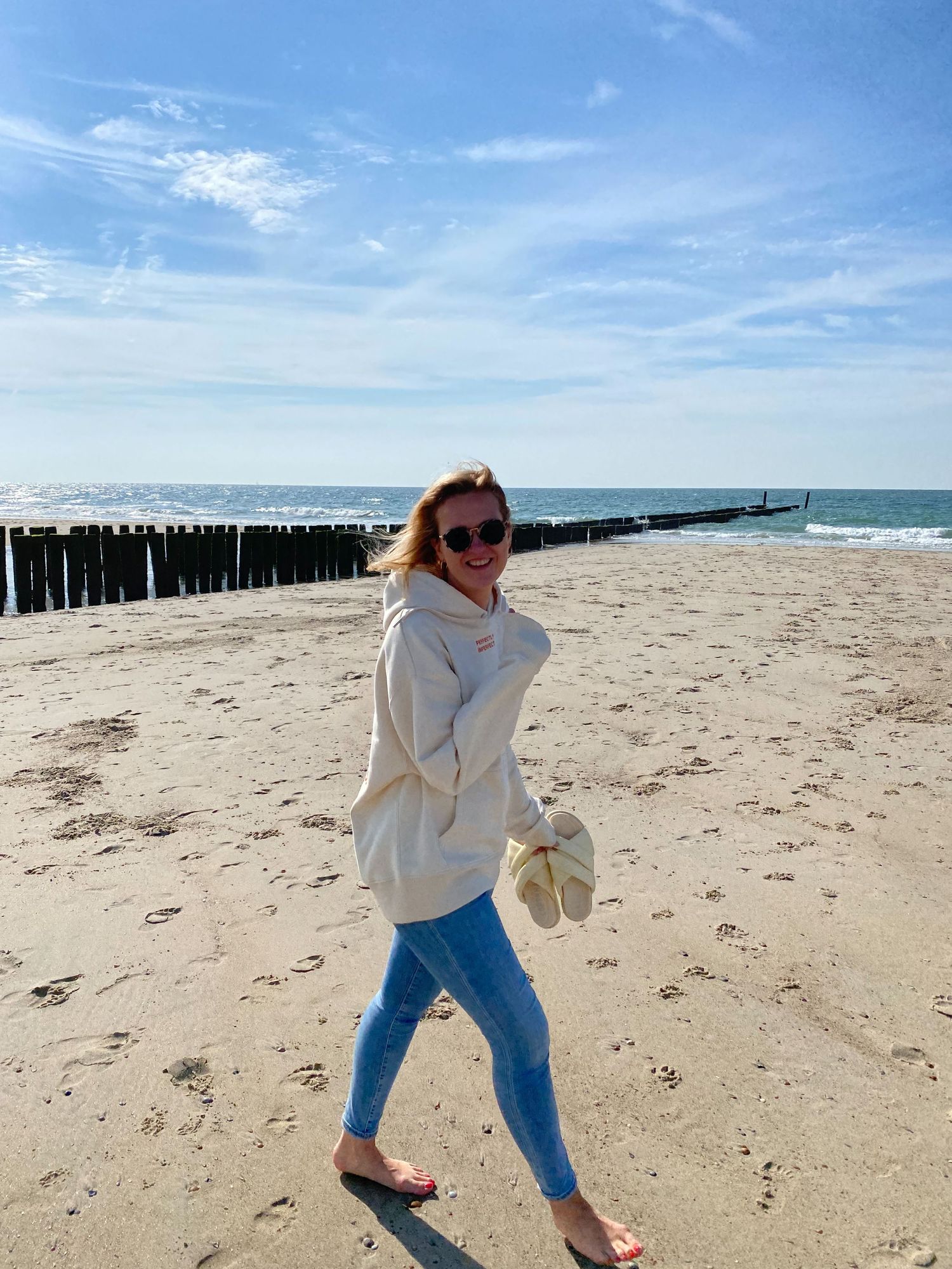 Person walking on beach with sunglasses and white sweater holding sandals near wooden pier.