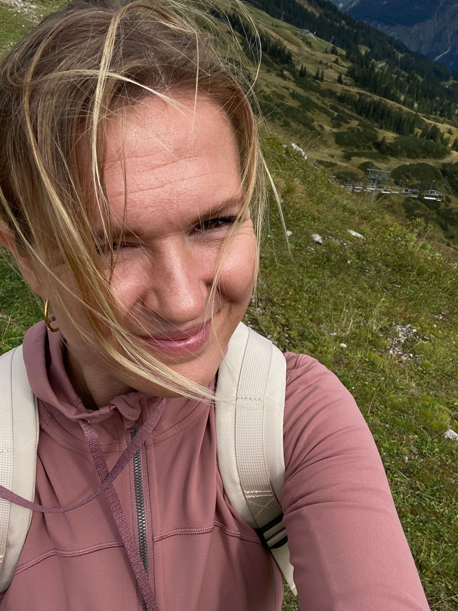 Person in pink athletic jacket smiling during mountain hike with scenic backdrop.