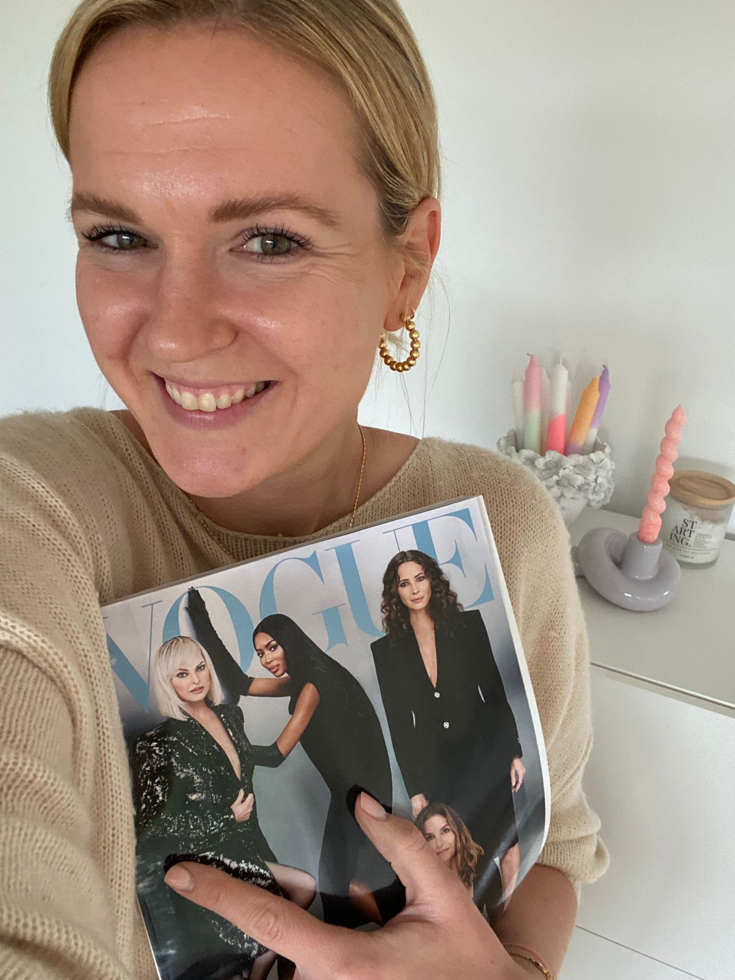 Person holding Vogue magazine while wearing beige sweater with candles in background.