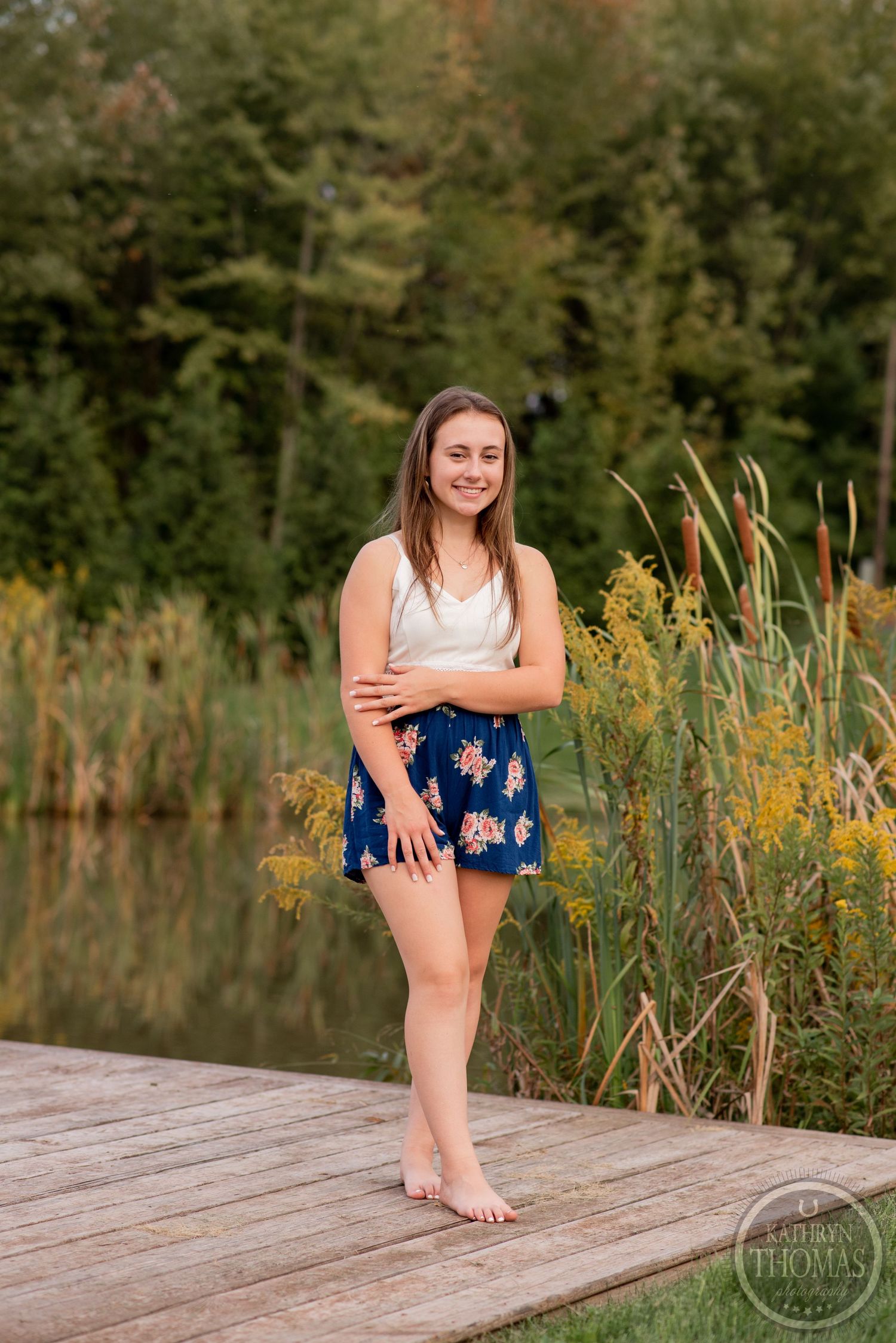 A person stands barefoot on a wooden dock surrounded by cattails and natural scenery.