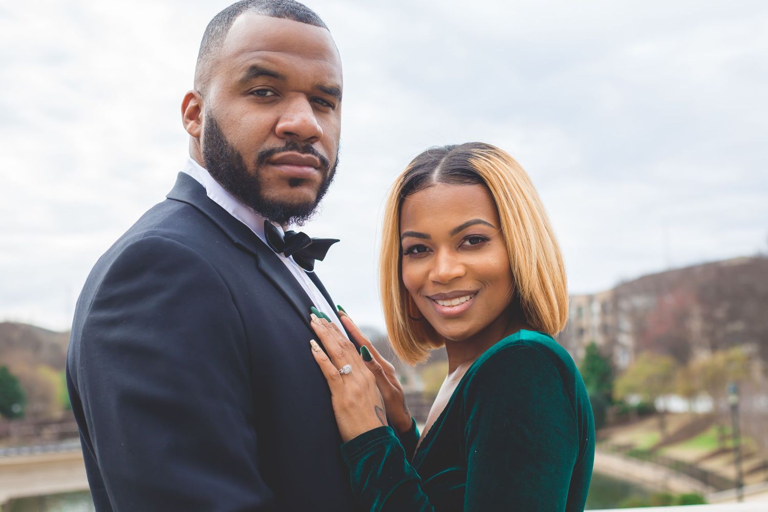A couple poses together outdoors, with one wearing a tuxedo and the other in an elegant green dress.