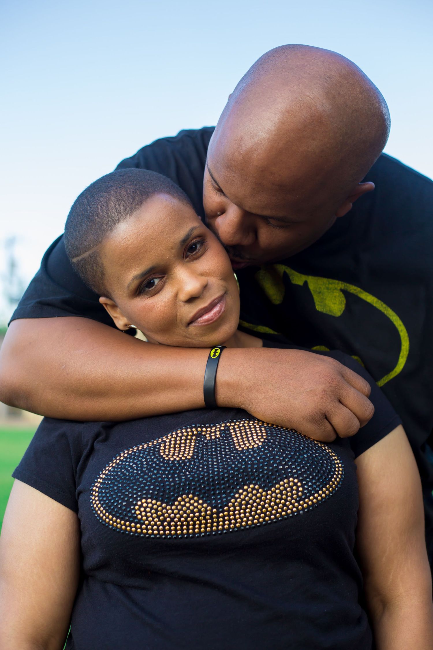 Loving embrace between parent and child wearing Batman shirt outdoors against blue sky.