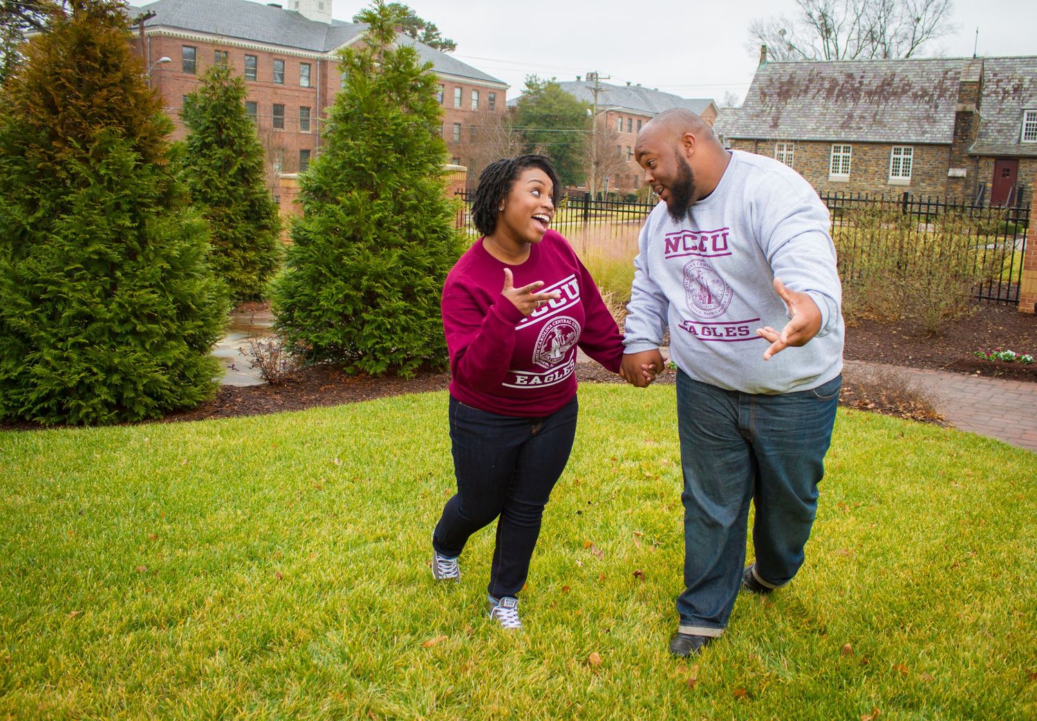 Playful couple laughing and dancing together on a grassy lawn with trees in background.