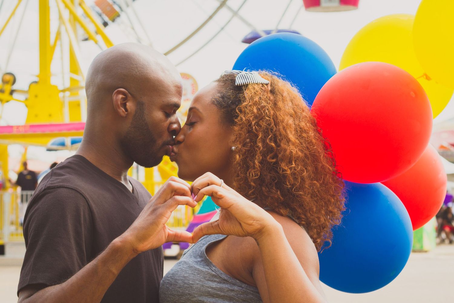A couple forms a heart shape with their hands against a backdrop of colorful carnival balloons.