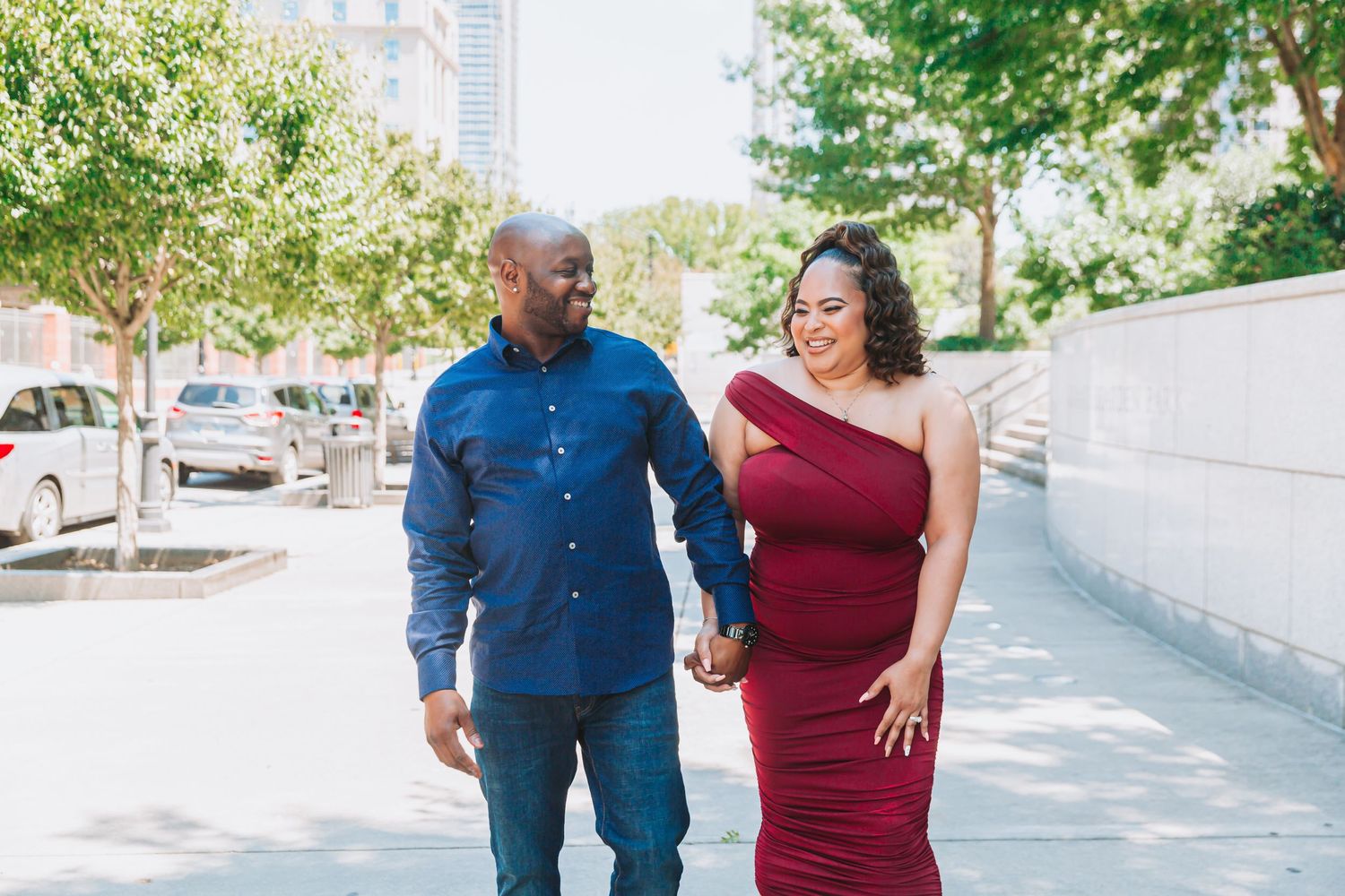 Couple walking together on sunny sidewalk, with woman in red dress and man in blue button-down shirt.