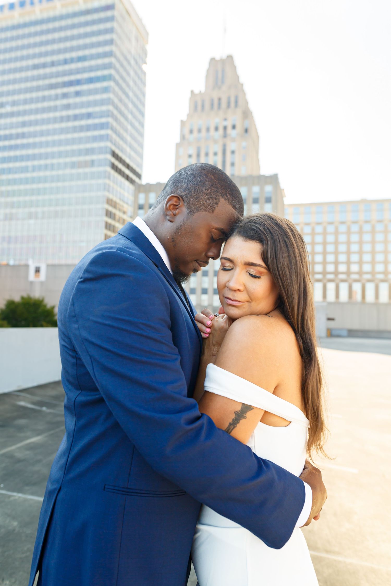 A romantic couple embraces on a rooftop with city buildings in the background at sunset.
