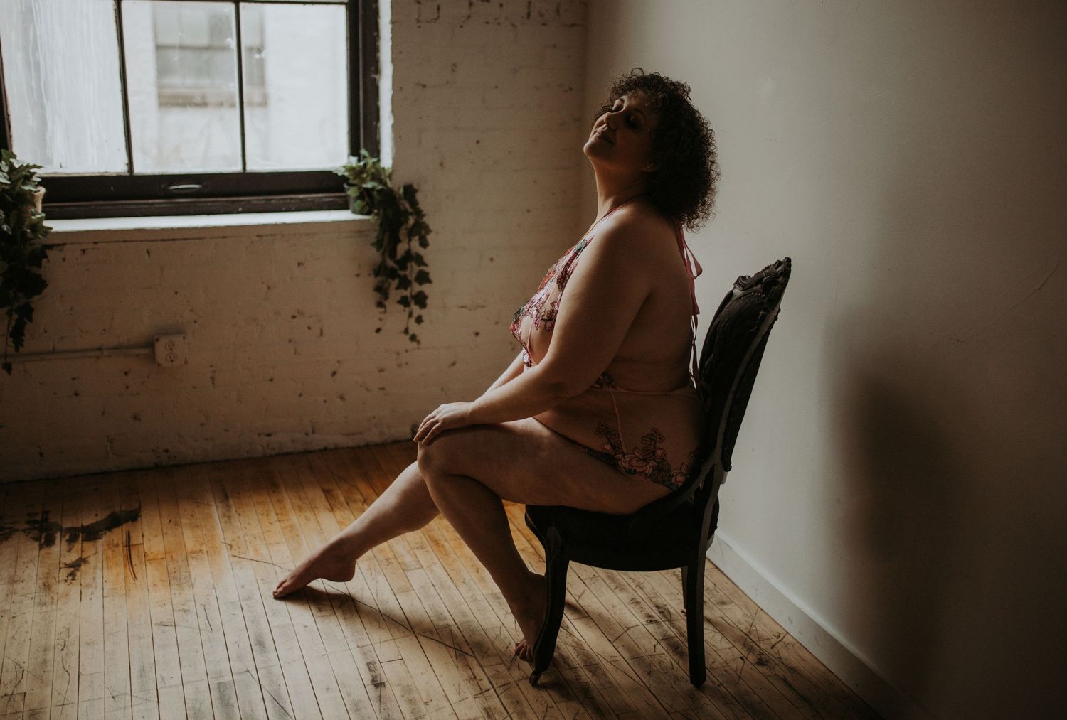 A person sits in profile on a black chair near a window with moody, natural light streaming into a minimalist room.
