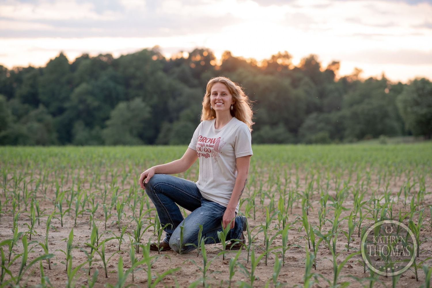 At sunset, figure kneels in emerging crop field with trees in background.