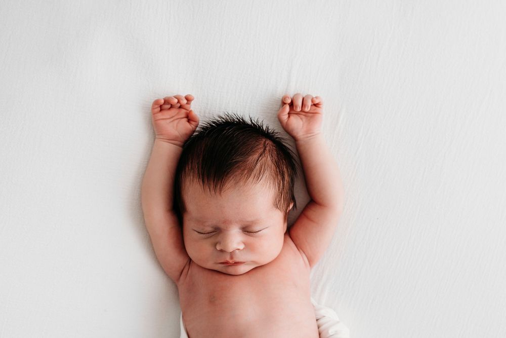 A sleeping newborn baby lies peacefully on a white blanket with arms raised overhead.