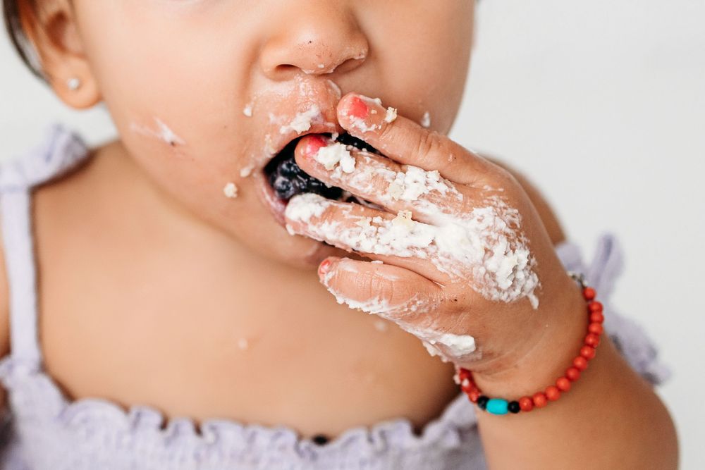 Close-up of baby wearing a colorful beaded bracelet messily eating cake with white frosting on their fingers and mouth.