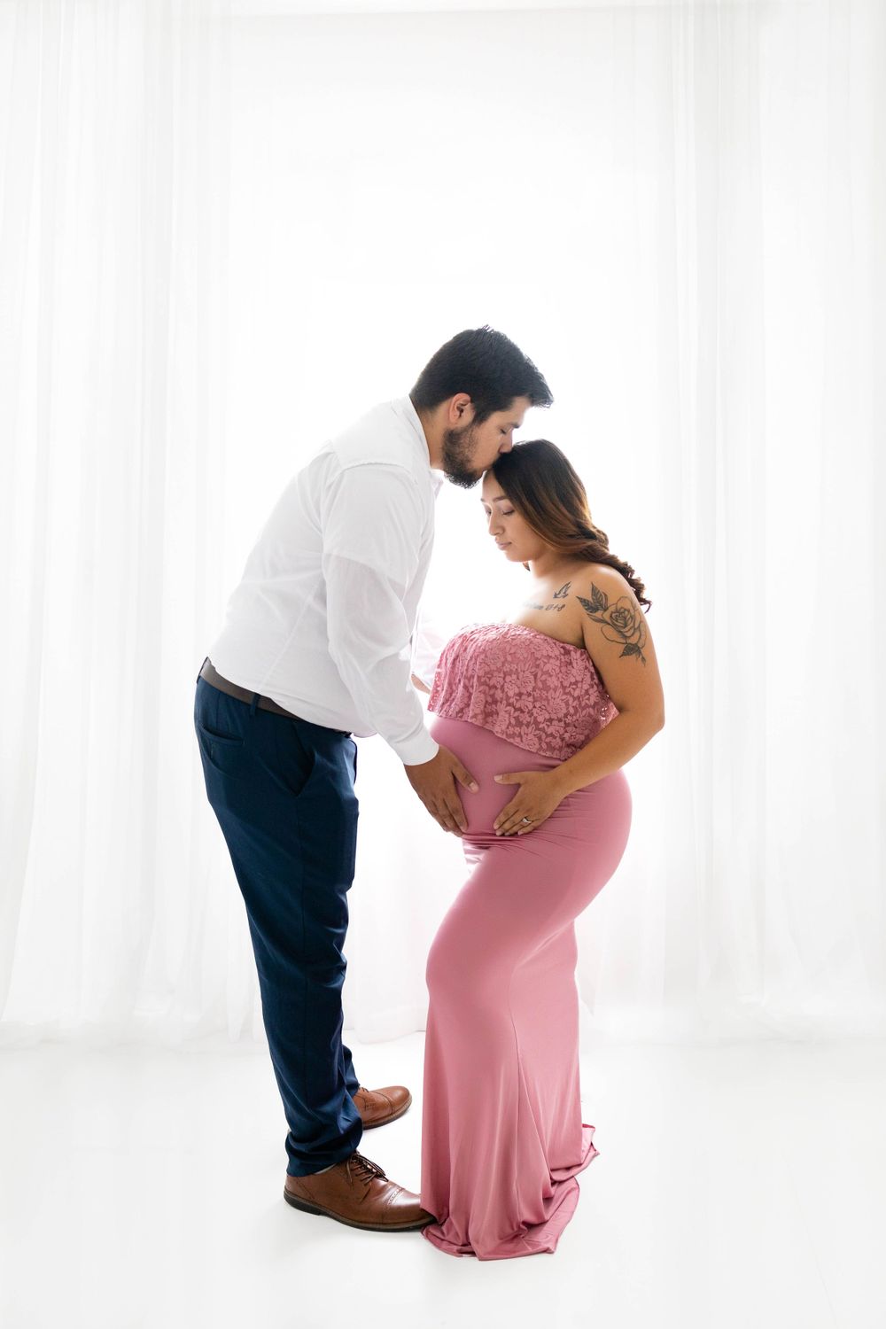Dad kissing pregnant mom-to-be's head and holding her baby belly in the minimalist white studio in Santa Fe NM. Dreamy lighting maternity.