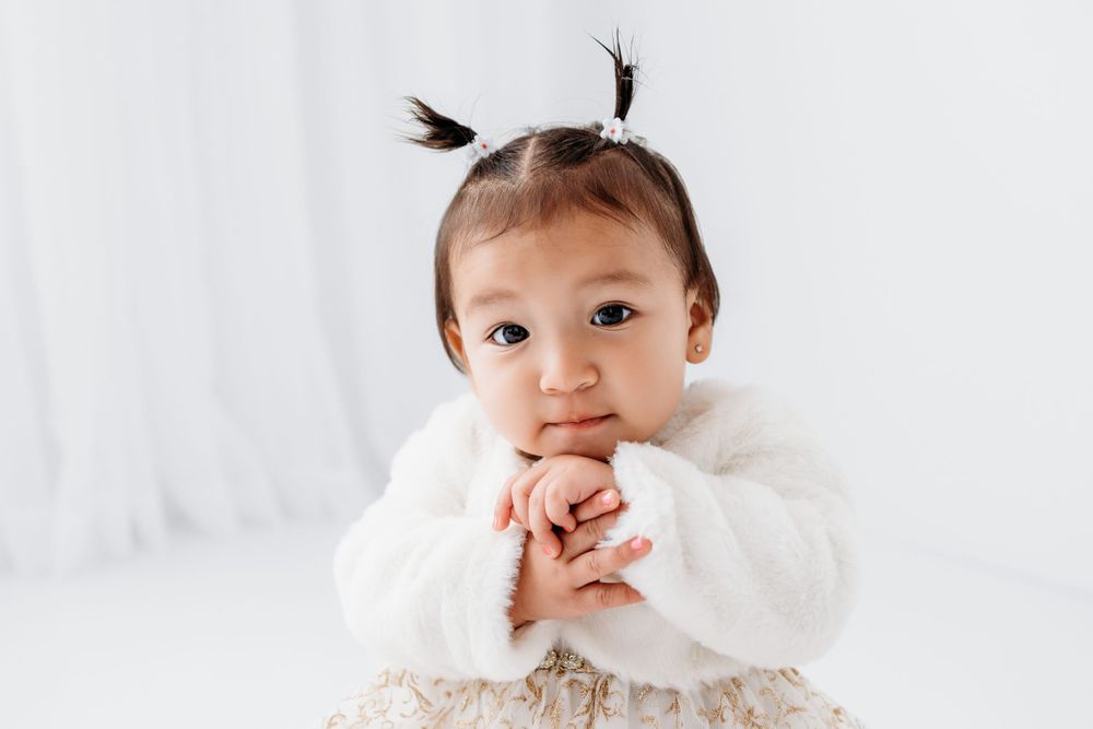 Baby in white dress smiles while sitting against light background wearing fur coat and wearing pigtails