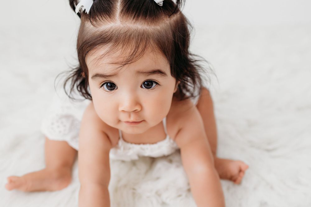 Baby in white lace dress crawls on soft white blanket during milestone photo session.