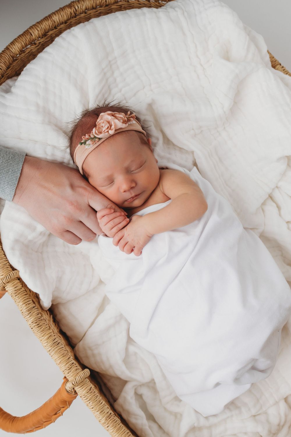 Newborn baby girl with floral headband in a Moses basket holding mom's finger at Blooming Desert Photography studio in Santa Fe New Mexico.