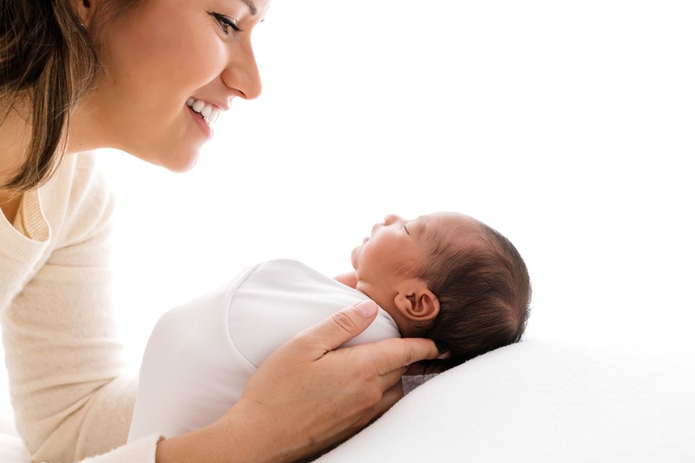 A tender moment between mom and newborn baby as they cuddle together in soft white clothing and swaddle