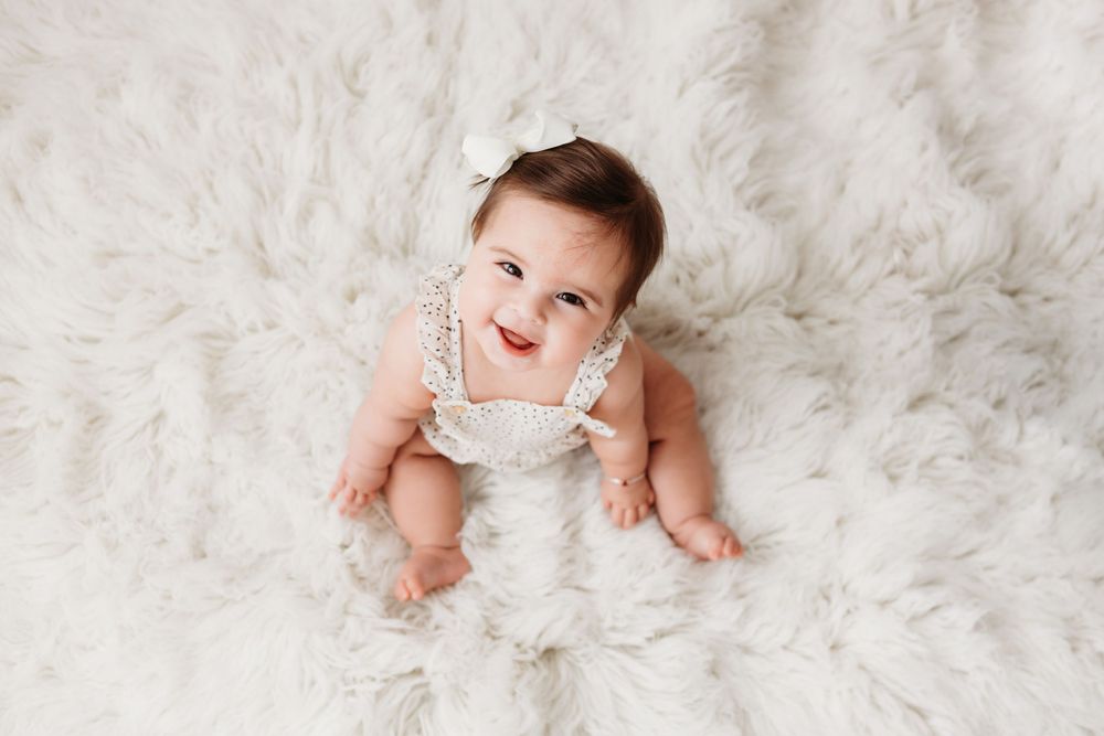 Baby wearing white outfit lies on soft white fur blanket while smiling at camera. She has dimples and white bow in her hair