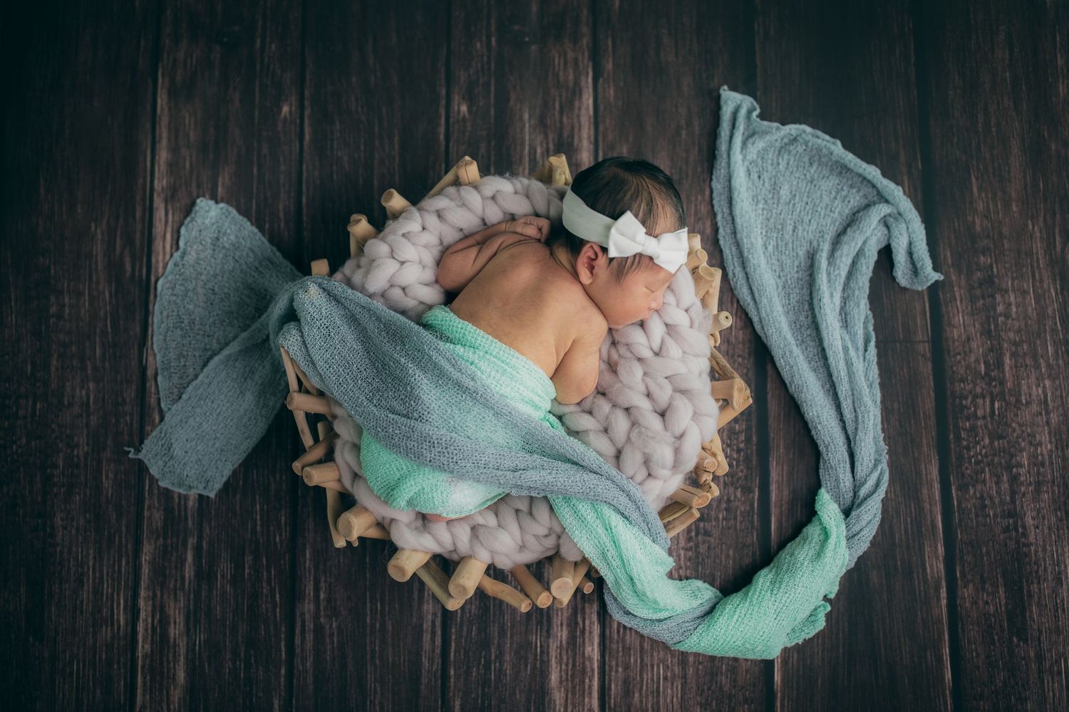 Newborn sleeping on a white blanket with mint green fabric wrapping on dark wooden floor.