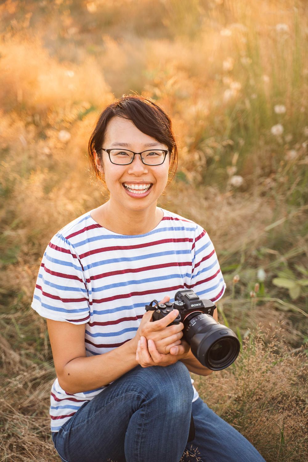 Seattle family photographer Ling Ling smiling and holding her Sony a7iv camera in a field backlit at golden hour