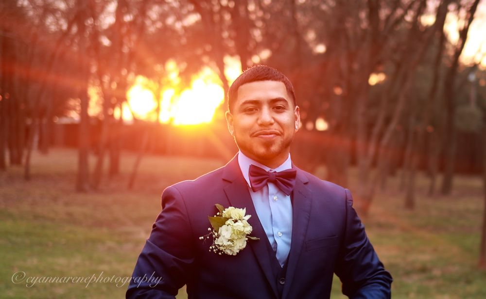 A well-dressed person in a navy suit with red bowtie and boutonniere smiles at sunset in a wooded setting.