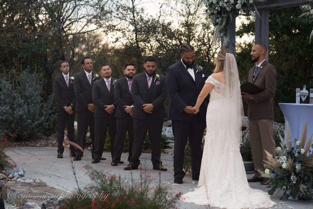 A wedding ceremony takes place outdoors at sunset with people gathered on a stone patio surrounded by greenery.