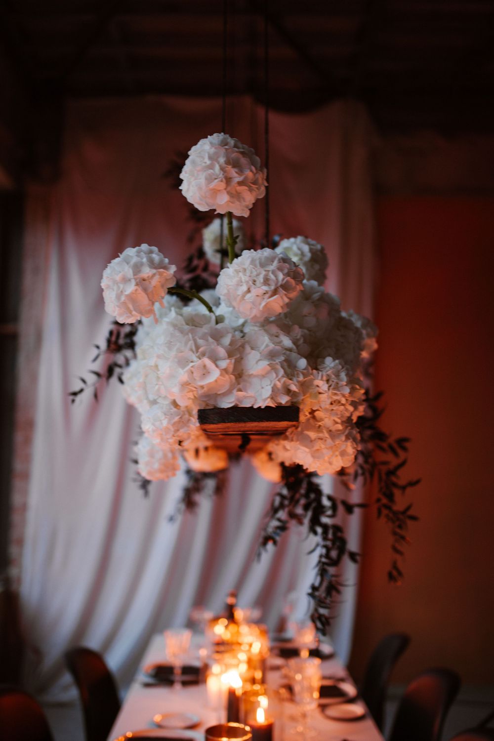 Elegant white hydrangea floral arrangements illuminated by warm candlelight against white draping.