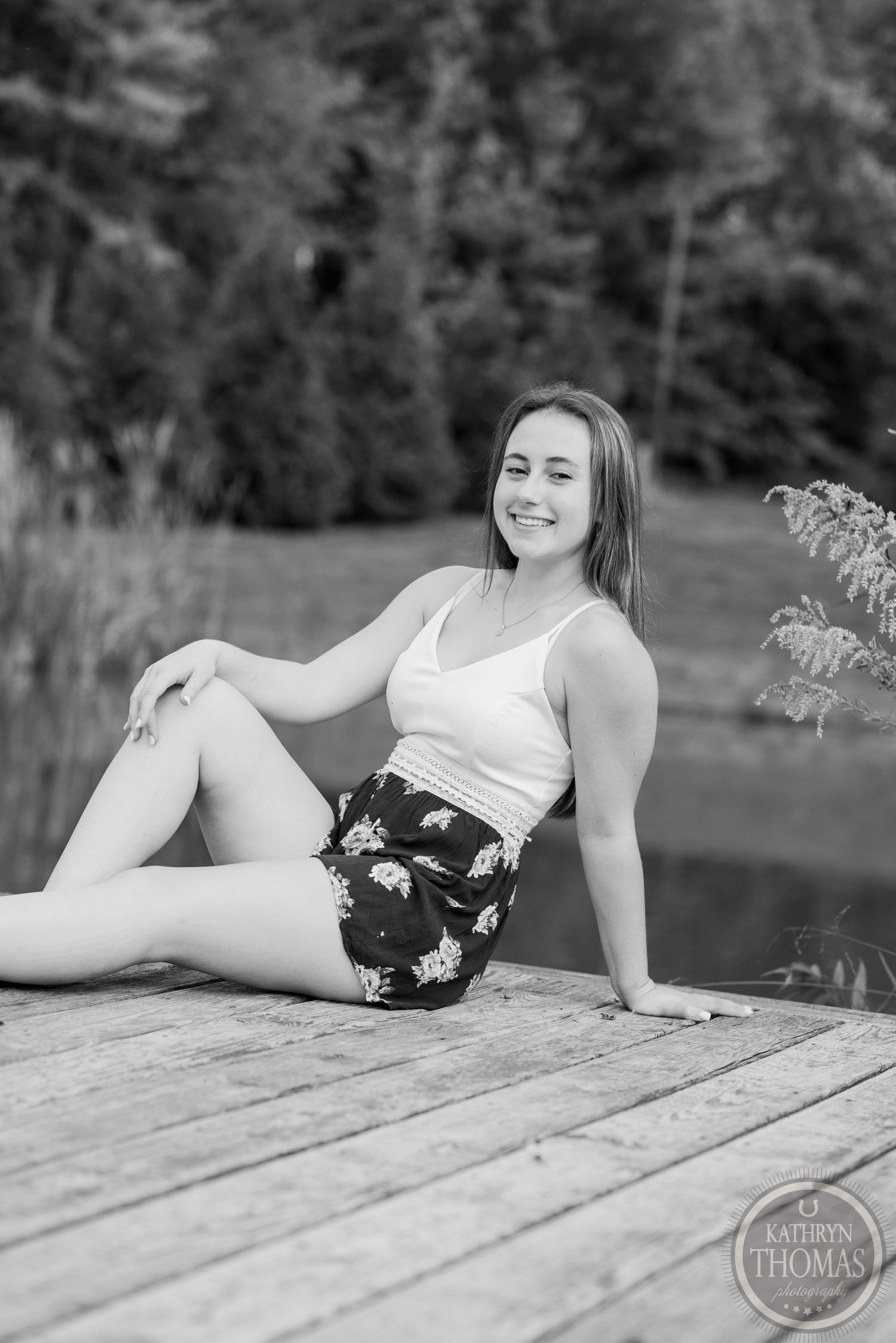 A black and white portrait of someone sitting casually on a wooden dock by a lake.