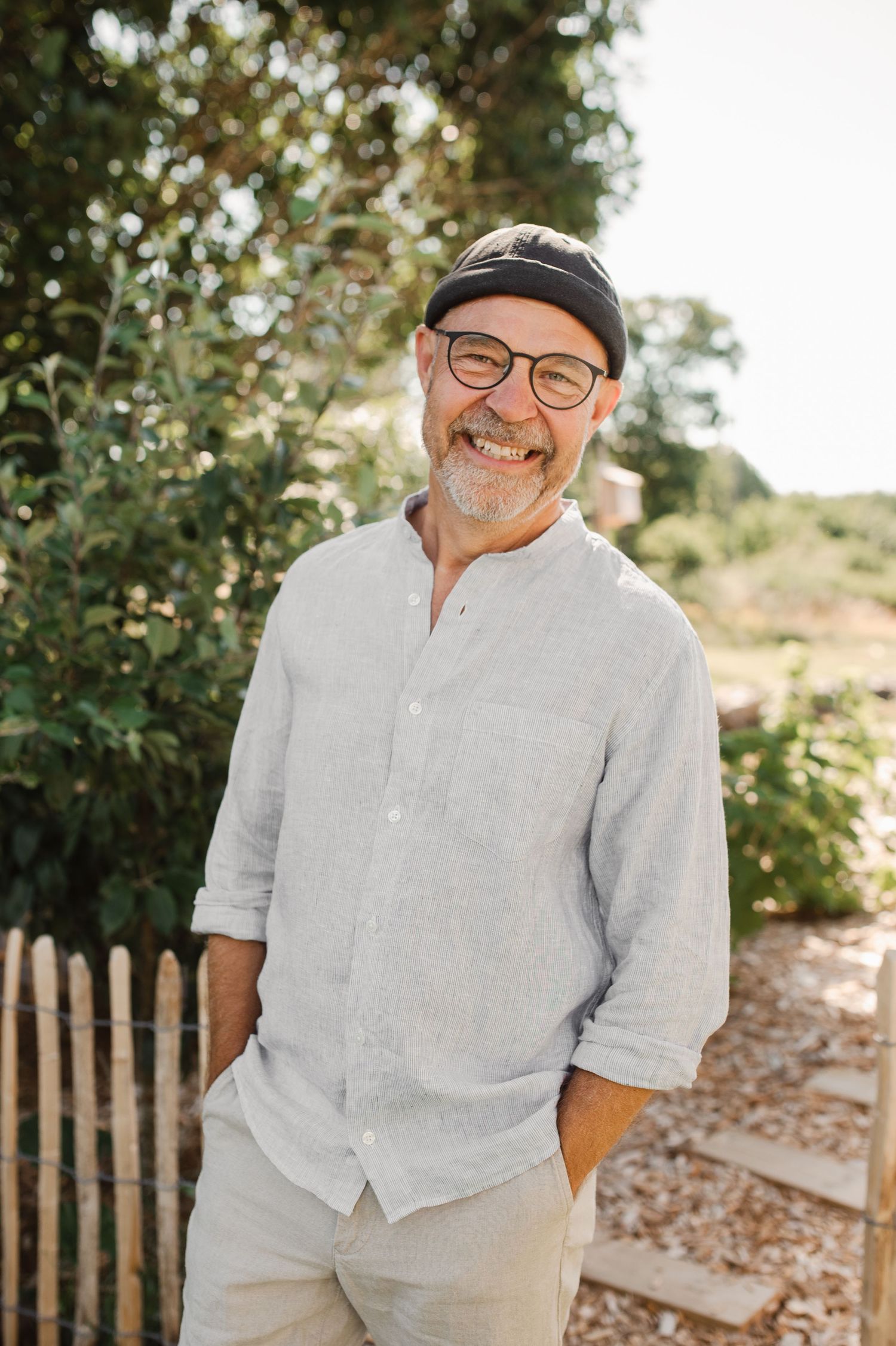 Person wearing glasses and casual linen shirt smiles warmly in outdoor garden setting.