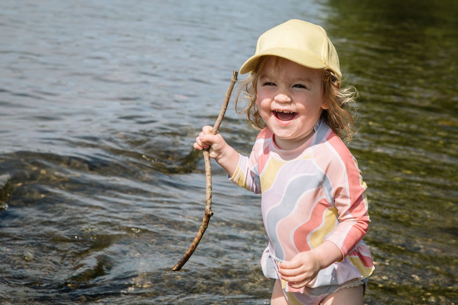 Child in sun protection gear holds a stick while playing by the water's edge.
